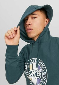 Sudadera verde con capucha ajustable, que presenta un gran gráfico circular en la parte frontal y puños acanalados. Textura suave.