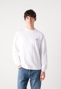 Sudadera blanca con cuello redondo, corte relajado y mangas largas. Presenta un pequeño logo azul en el pecho. Combinada con jeans azules.