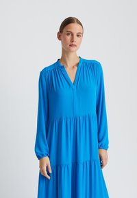 Femme aux cheveux attachés portant une robe longue à manches longues, de couleur bleu vif, à volants et col en V, debout devant un fond uni.