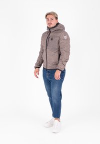 Giubbotto trapuntato marrone con cappuccio, zip frontale e dettagli neri; abbinato a jeans blu e sneakers bianche.