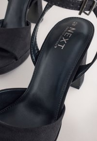 Sorte ruskindssandaler med høje hæle og en robust hæl, med åben tå og ankelrem, der fremhæver en glat læderfodflade.