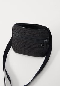 Černá crossbody taška s hladkým povrchem, která má opakující se vzor loga, zipové přihrádky a pevný textilní popruh.