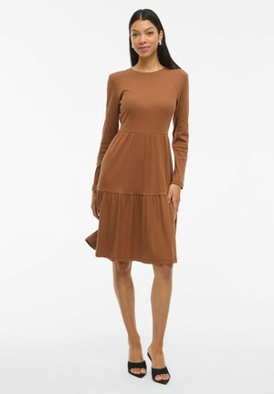 Robe marron à manches longues en matière côtelée, avec une jupe à volants et un col rond. Elle a une coupe décontractée et une longueur mi-mollet.
