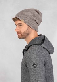Grijze gebreide beanie met een slouchy ontwerp, met ribbeltextuur en een klein logo-label. Draagt een grijze hoodie met een trekkoordcapuchon.
