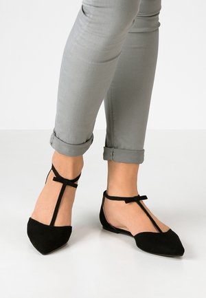 Black suede flat shoes with a pointed toe, featuring a T-strap design and a delicate ankle tie. The model is wearing light gray rolled jeans.