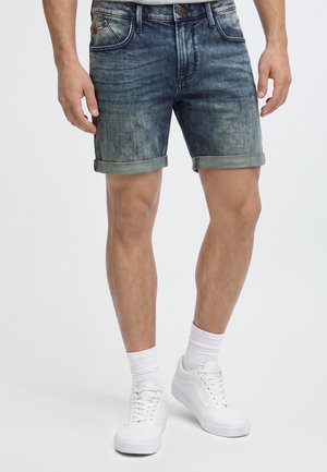 Homme portant un short en denim bleu foncé retroussé, des chaussettes blanches et des baskets blanches, debout sur un fond clair uni.