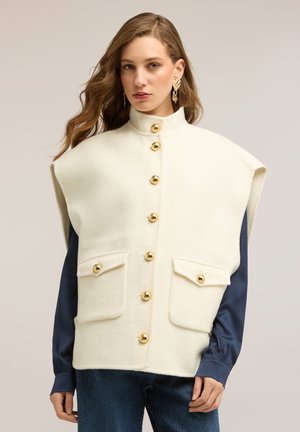 Donna con capelli lunghi e mossi indossa un gilet bianco senza maniche con bottoni dorati e tasche sopra una camicia blu scuro a maniche lunghe su sfondo neutro.