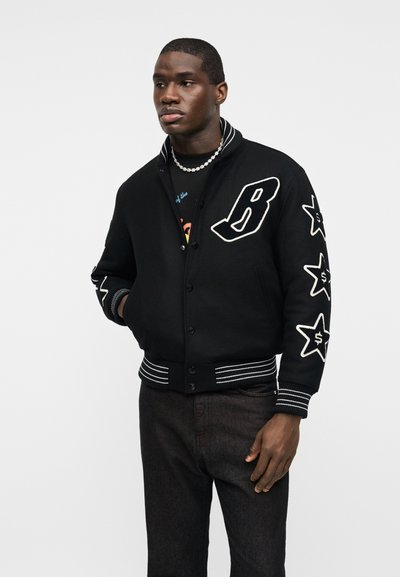 Άνδρας που φοράει μαύρο βαρσιβαλικό μπουφάν με μεγάλο γράμμα "B" στο στήθος και λευκά αστέρια ραμμένα στα μανίκια, συνδυασμένα με σκουρόχρωμο παντελόνι και κολιέ με χάντρες.