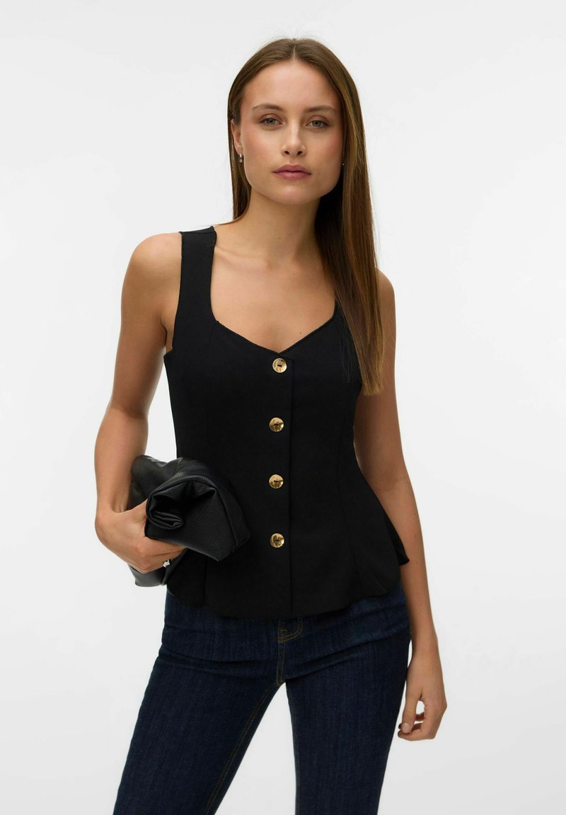 Jeune femme portant un haut noir sans manches à boutons et un jean foncé, tenant une pochette noire pliée devant un fond uni.