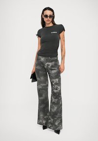T-shirt noir ajusté avec un logo blanc, associé à un pantalon flare grigio camouflage taille haute. Le mannequin tient une pochette noire et porte des lunettes de soleil.