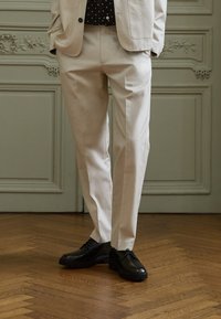 Beige byxor med skräddarsydd passform och slät textur, kombinerade med svarta snörskor på ett trägolv.