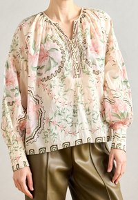 Femme portant un chemisier crème à motif floral rose et vert avec des manches bouffantes, associé à un pantalon plissé vert olive.