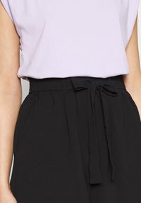 Woman wearing a light purple sleeveless top tucked into high-waisted black pants with a tied fabric belt at the waist.