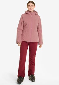 Rosa wasserdichte Jacke mit Kapuze, Frontreißverschluss und Seitentaschen, kombiniert mit figurbetonten burgunderroten Hosen und schwarzen Skischuhen. Glatte Textur.