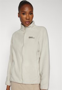 Giacca in pile beige chiaro con colletto alto, zip completa e dettagli del logo sul lato sinistro del petto. Tessuto morbido e maniche lunghe.