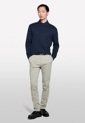 Pull en maille navy, chinos beiges, ceinture noire et chaussures noires. Le pull présente un col montant et une coupe décontractée. Design simple et épuré.