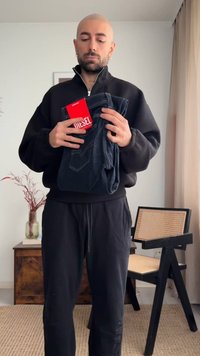 Glatzköpfiger Mann in schwarzem Sweatshirt und Hose hält zusammengefaltete Diesel-Jeans mit rotem Etikett und steht in einem minimalistisch eingerichteten Raum mit einem Stuhl und einer Vase in der Nähe.