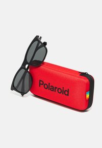 Zwarte zonnebril die rust op een rode rechthoekige Polaroidhoes met een zwarte ritssluiting en een klein regenbooglabel aan de zijkant.