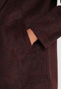 Vinrød uldfrakke med en blød struktur, der har en sidelomme og en ren, skræddersyet silhuet. Stoffet har subtile variationer i farvetonen.