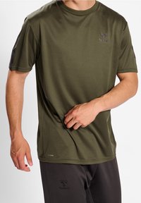 Man i olivgrön t-shirt med kort ärm och svarta träningsbyxor, som justerar skjortan vid midjan.