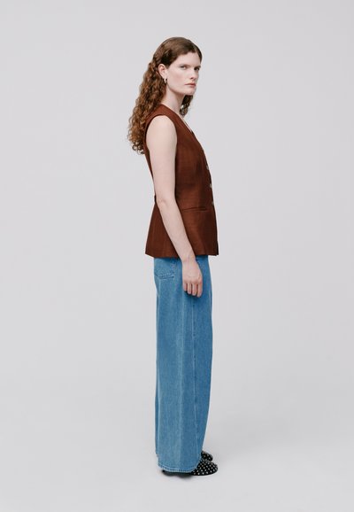 Gilet senza maniche in lino marrone con una forma aderente, abbinato a pantaloni in denim blu a gamba larga e scarpe nere con pois bianchi.