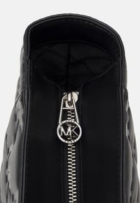 Sac à main noir avec fermeture éclair, doté d'un logo rond en argent MK sur le tirette de la fermeture, extérieur en cuir texturé et doublure en tissu lisse.