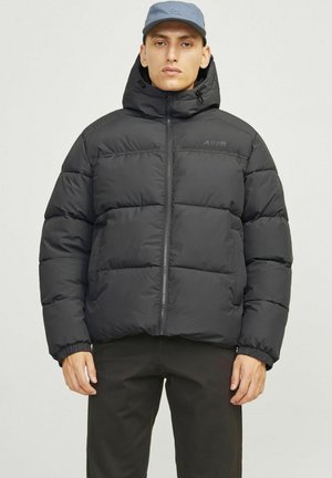 Chaqueta puffer negra con capucha, que presenta un cierre frontal, puños elásticos y textura acolchada. Combinada con una gorra gris.
