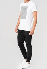 White cotton t-shirt featuring a black graphic design, paired with black tapered pants and white sneakers with a textured surface.