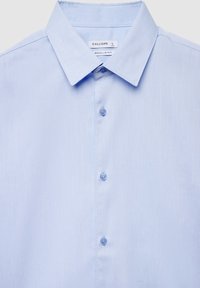 Hellblaues, regulär geschnittenes Hemd mit strukturierter Musterung, spitzem Kragen und sechs passenden Knöpfen entlang der Vorderseite. Das Etikett trägt die Aufschrift "CALLIOPE."