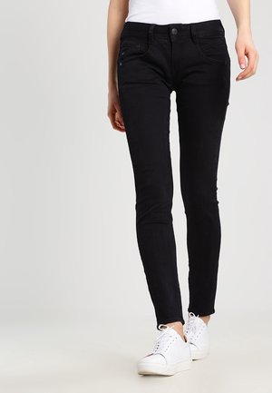 Personne portant un jean noir slim et des baskets blanches, debout devant un fond gris clair uni.