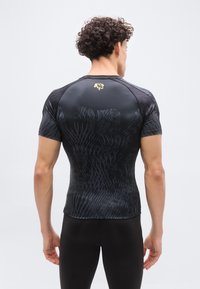 Νεαρός άνδρας με σγουρά μαλλιά, φοράει στενό αθλητικό μπλουζάκι με μαύρα σχέδια και μαύρα κολάν, στέκεται με την πλάτη προς τα εμπρός σε απλό φόντο.