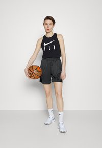 Zwarte mouwloze tanktop met de tekst "FLY", gecombineerd met zwarte shorts. Houdt een oranje basketbal vast; draagt witte sokken en lichtgrijze sneakers.