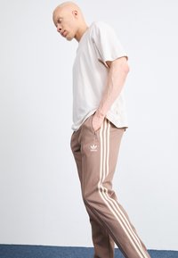 Lichtbeige t-shirt en bruine trainingsbroek met witte strepen. De broek heeft een ritsdetail en een Adidas-logo op de linker bovenbeen.