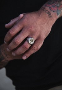 Silverring med en cirkulär platta som har ett palmträdsmotiv; intrikat textur med gulddetaljer; bärs på en hand med synliga tatueringar.