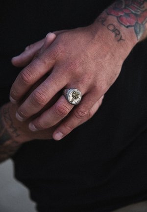 Bague en argent avec un visage circulaire présentant un motif de palmier ; texture intricate avec des accents dorés ; portée sur une main avec des tatouages visibles.