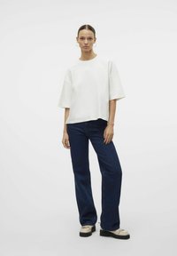 Bílý oversized top s krátkým rukávem, který je zkombinován s tmavě modrými širokými džíny a béžovými platformovými botami. Minimalistický design, hladká textura.