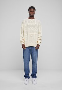 Chunky kremfarget strikkegenser med kabelmønster, kombinert med lyseblå denimjeans og hvite joggesko. Avslappet passform og lange ermer.