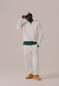 Cremefarbener Reißverschluss-Hoodie über einem grünen Shirt, kombiniert mit passenden cremefarbenen Jogginghosen und braunen Schuhen. Das Outfit hat eine lässige Passform.