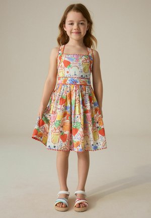 Fiatal lány, aki színes ujjatlan ruhát visel gyümölcs- és virágmintákkal, valamint fehér szandált többszínű pántokkal.