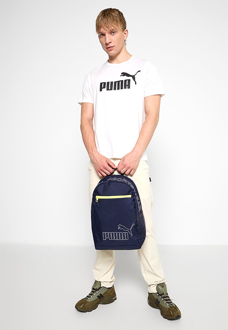 Joven vestido con una camiseta blanca de Puma y pantalones beis, sosteniendo una mochila navy de Puma con cremallera amarilla, de pie frente a un fondo blanco.
