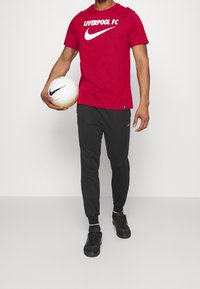 Punainen puuvillapaita, jossa on valkoiset tekstit "LIVERPOOL FC" ja Nike-logo, yhdistetty mustiin kapeisiin verkkareihin ja mustiin urheilukenkihin, pitäen valkoista jalkapalloa.