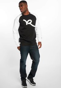 Felpa in bianco e nero con un grande logo bianco, abbinata a jeans blu scuro e sneakers nere. La felpa ha un design a maniche raglan.