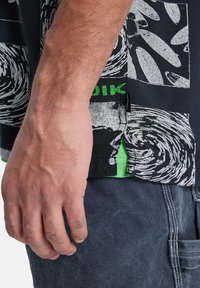 Κοντομάνικο πουκάμισο σε μαύρο χρώμα με λευκό σχέδιο σπειροειδούς μορφής, πράσινες λεπτομέρειες και πλαϊνή σχισμή. Το ύφασμα φαίνεται λείο και ελαφρύ.