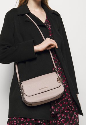 Mujer con abrigo negro y vestido floral sosteniendo un bolso bandolera Michael Kors rosa claro con colgante de logo dorado.