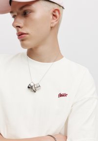 Hvid T-shirt med rødt broderet logo, båret med en sølvvedhæng halskæde, der fremhæver det minimalistiske design. Pink cap er synlig.