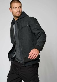 Veste noire en texture avec une capuche, dotée d'une fermeture éclair frontale et de poches latérales. Portée sur un sweat-shirt gris, complétée par un pantalon sombre.