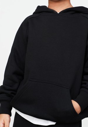 Enfant portant un sweat à capuche noir avec une poche frontale, un t-shirt blanc visible en dessous, les mains partiellement dans les poches.