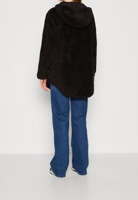 Persona che indossa una giacca di pile con cappuccio nera e jeans blu, in piedi di spalle su uno sfondo bianco semplice.