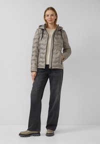 Vrouw staat tegen een effen achtergrond gekleed in een lichtgrijze gewatteerde hoodie, een crèmekleurige trui, donker lange broek en grijze laarzen.