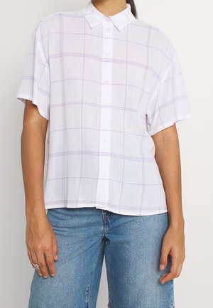 Person wearing a white short-sleeve button-up shirt with light purple plaid pattern and blue jeans, standing against a plain background.
